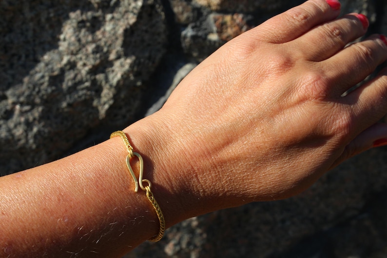 May include: A gold-toned bracelet featuring a fine, textured chain and a prominent hook-and-eye style clasp mechanism. The bracelet is crafted with a matte finish and displays a consistent, intricate woven pattern throughout the entire length of the band.
