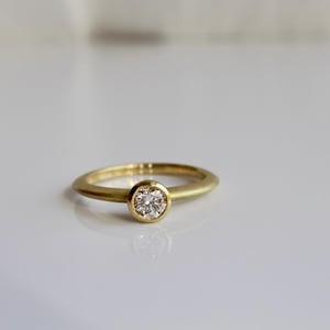 May include: A gold ring with a single, round, clear diamond set in a bezel. The ring band is a simple, smooth design. The ring is displayed on a white surface.