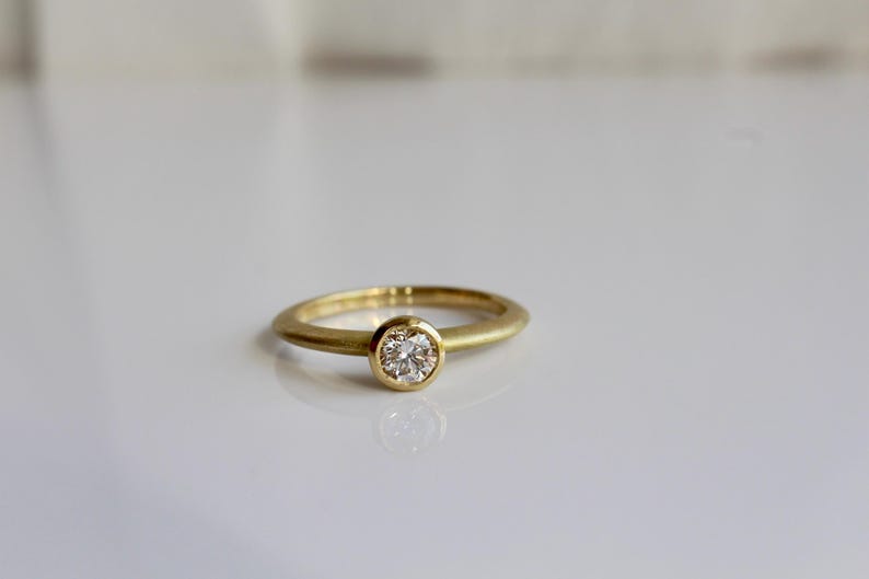 May include: A gold ring with a single, round, clear diamond set in a bezel. The ring band is a simple, smooth design. The ring is displayed on a white surface.