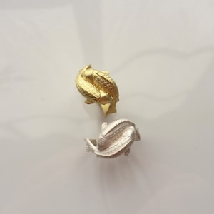 May include: Two rings, one gold-colored and one silver-colored, each featuring a design of two fish intertwined. The rings are displayed on a white surface, showcasing their intricate details and metallic sheen.