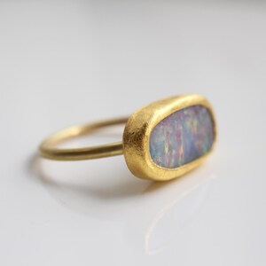 May include: A gold ring featuring an oval-shaped opal gemstone with iridescent flashes of blue, pink, and green. The ring has a textured, matte gold finish on the bezel setting and a smooth, thin, rounded gold band that connects to the side.