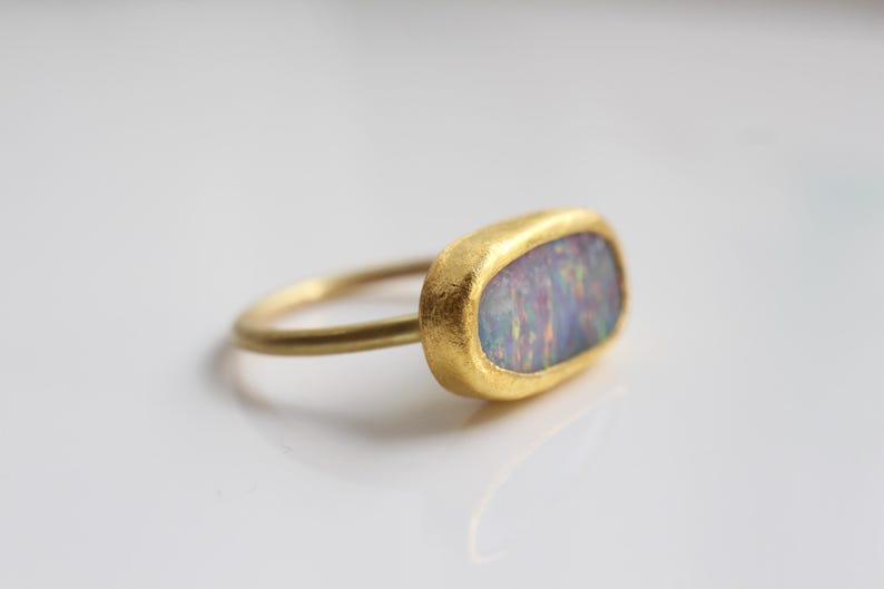 May include: A gold ring featuring an oval-shaped opal gemstone with iridescent flashes of blue, pink, and green. The ring has a textured, matte gold finish on the bezel setting and a smooth, thin, rounded gold band that connects to the side.