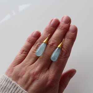 May include: A pair of teardrop-shaped earrings with pale blue gemstones and gold-colored metal accents. The earrings are displayed on a hand, showcasing their delicate design and color. The jewelry is a stylish accessory.
