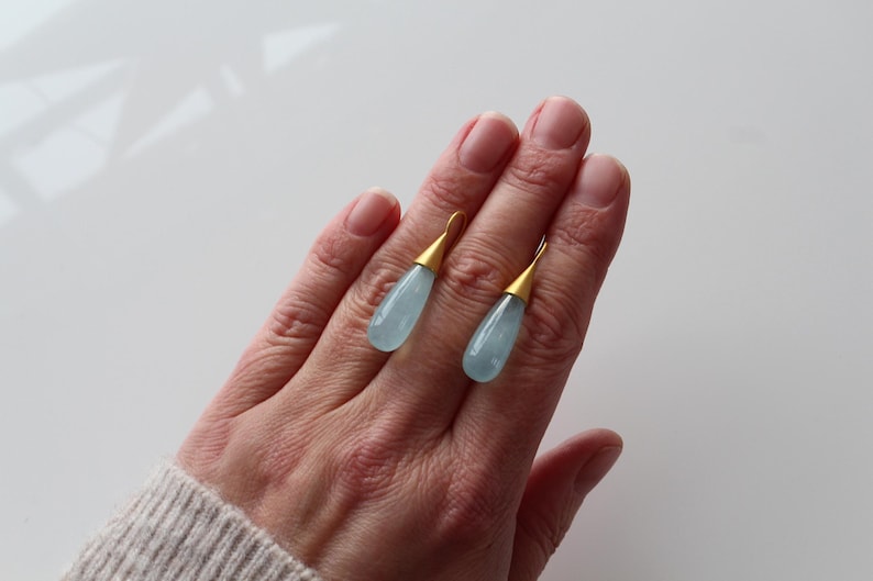 May include: A pair of teardrop-shaped earrings with pale blue gemstones and gold-colored metal accents. The earrings are displayed on a hand, showcasing their delicate design and color. The jewelry is a stylish accessory.