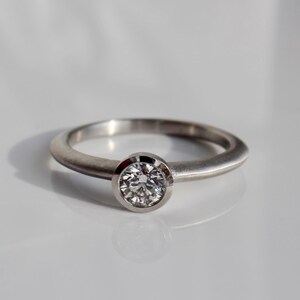 May include: A silver ring with a round, clear gemstone set in a bezel. The ring band is smooth and polished. The ring is displayed on a white surface, with a shadow cast to the side.