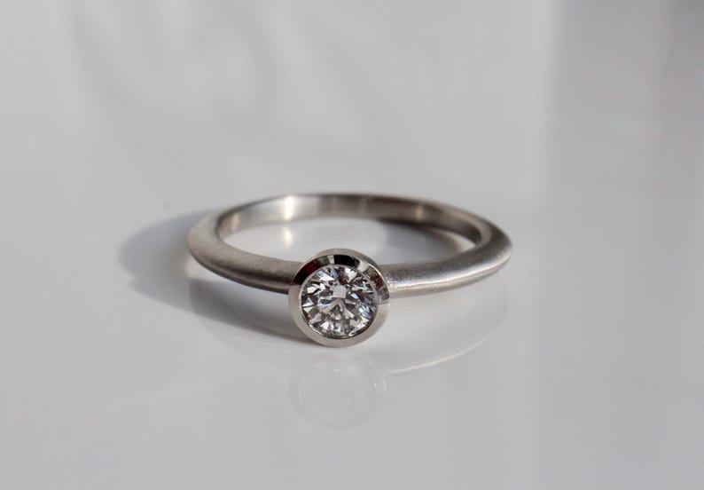 May include: A silver ring with a round, clear gemstone set in a bezel. The ring band is smooth and polished. The ring is displayed on a white surface, with a shadow cast to the side.
