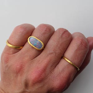 May include: A set of three gold rings, with the center ring featuring an oval-shaped opal gemstone displaying iridescent flashes of blue, pink, and green. The rings have a smooth, matte gold finish and a simple, rounded band design.