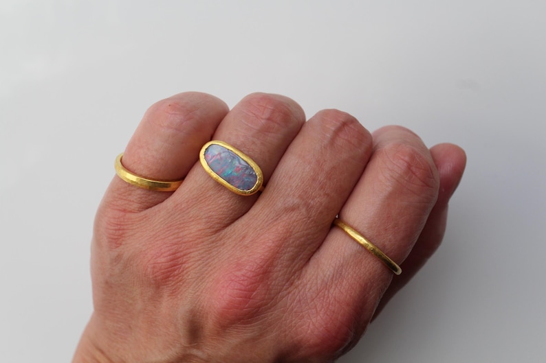 May include: A set of three gold rings, with the center ring featuring an oval-shaped opal gemstone displaying iridescent flashes of blue, pink, and green. The rings have a smooth, matte gold finish and a simple, rounded band design.