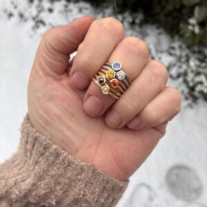 May include: A stack of five thin metal rings worn on a finger. Each ring features a small, round, bezel-set gemstone in a different color, including light blue, yellow, clear, orange, and dark brown, set in silver, gold, and rose gold bands.