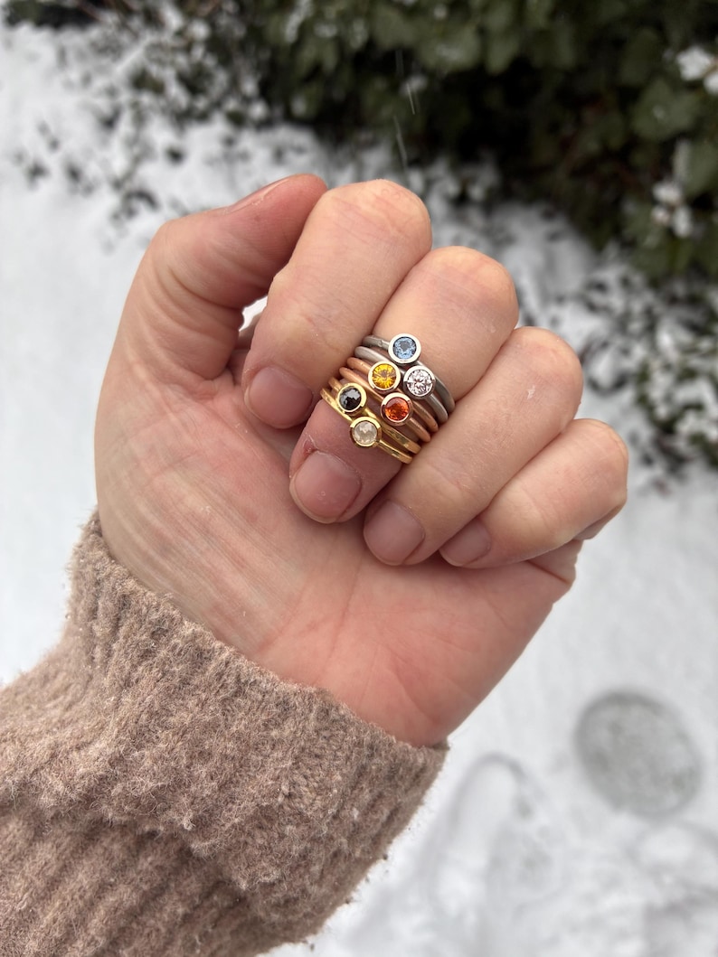 May include: A stack of five thin metal rings worn on a finger. Each ring features a small, round, bezel-set gemstone in a different color, including light blue, yellow, clear, orange, and dark brown, set in silver, gold, and rose gold bands.