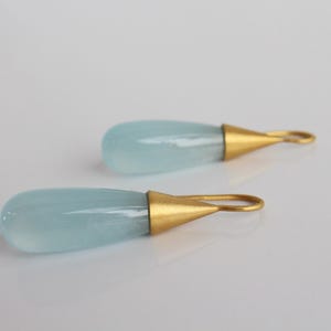 May include: A pair of teardrop-shaped earrings. Each earring features a pale blue, translucent stone with a gold-colored metal cap and loop. The earrings are displayed on a white surface.
