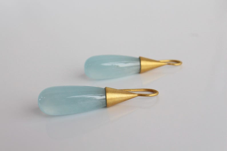 May include: A pair of teardrop-shaped earrings. Each earring features a pale blue, translucent stone with a gold-colored metal cap and loop. The earrings are displayed on a white surface.