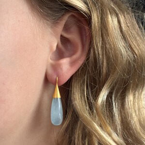 May include: Gold-tone drop earrings with a teardrop-shaped, pale blue gemstone. The earrings have a simple, elegant design, with a gold cap and hook. The gemstone is smooth and polished, reflecting light. The earrings are a classic accessory.