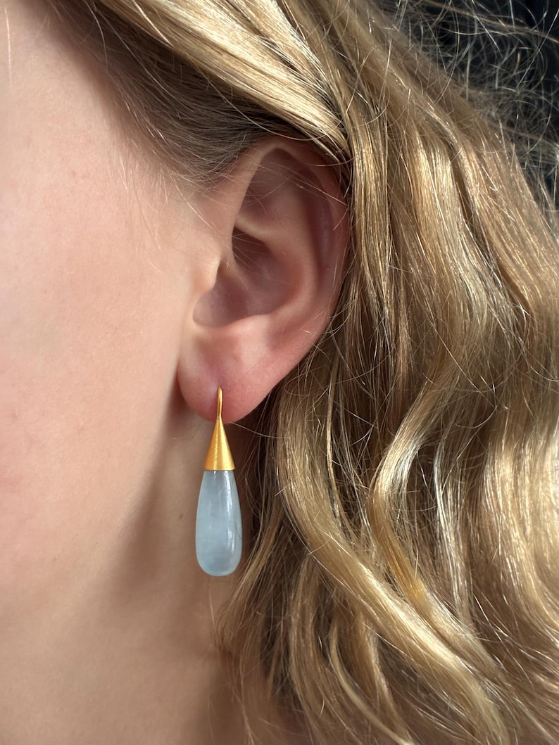 May include: Gold-tone drop earrings with a teardrop-shaped, pale blue gemstone. The earrings have a simple, elegant design, with a gold cap and hook. The gemstone is smooth and polished, reflecting light. The earrings are a classic accessory.