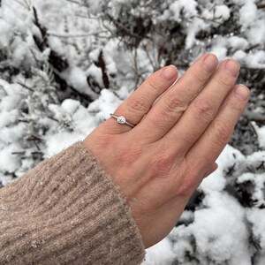 May include: A silver-toned ring featuring a single, round, clear gemstone set in a bezel setting. The band is thin and smooth, encircling the finger with a minimalist design that highlights the central stone's clarity and reflective surface.
