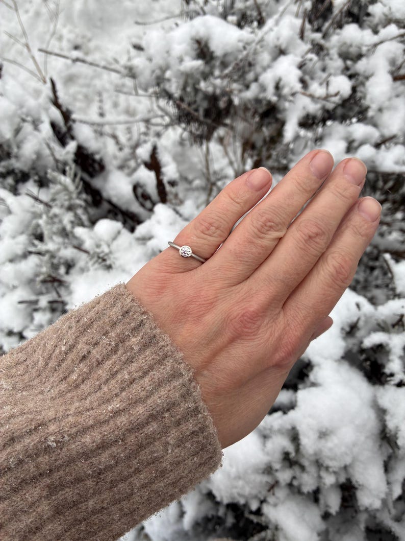 May include: A silver-toned ring featuring a single, round, clear gemstone set in a bezel setting. The band is thin and smooth, encircling the finger with a minimalist design that highlights the central stone's clarity and reflective surface.