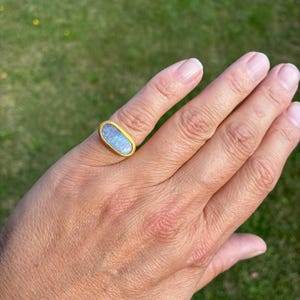 May include: A gold ring featuring an elongated oval-shaped opal gemstone. The opal displays iridescent flashes of blue, green, and pink colors against a milky white base. The metal band has a textured, matte gold finish surrounding the stone.