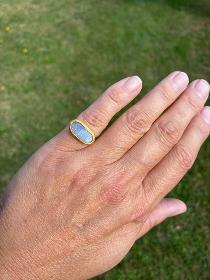 May include: A gold ring featuring an elongated oval-shaped opal gemstone. The opal displays iridescent flashes of blue, green, and pink colors against a milky white base. The metal band has a textured, matte gold finish surrounding the stone.