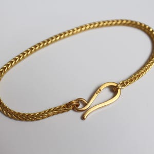 May include: A gold-colored bracelet featuring a woven wheat chain design. The bracelet is secured with a curved hook-style clasp. The metal surface has a polished finish, and the clasp is marked with a small stamp indicating its metal purity.