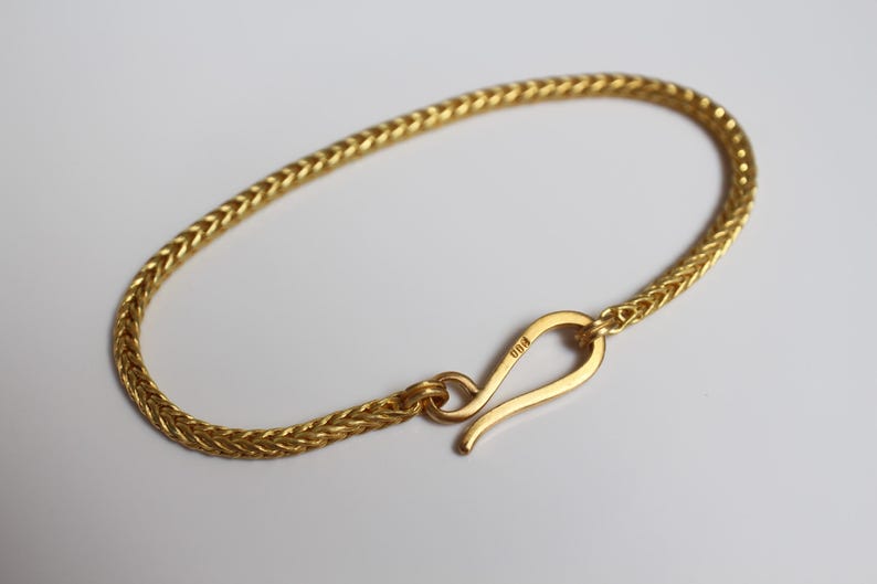 May include: A gold-colored bracelet featuring a woven wheat chain design. The bracelet is secured with a curved hook-style clasp. The metal surface has a polished finish, and the clasp is marked with a small stamp indicating its metal purity.