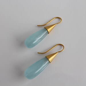 May include: A pair of elegant drop earrings. Each earring features a teardrop-shaped, pale blue gemstone suspended from a gold-colored hook. The gemstone is set in a gold-colored conical cap. The earrings are displayed on a white surface.