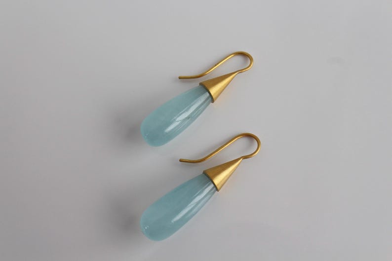 May include: A pair of elegant drop earrings. Each earring features a teardrop-shaped, pale blue gemstone suspended from a gold-colored hook. The gemstone is set in a gold-colored conical cap. The earrings are displayed on a white surface.