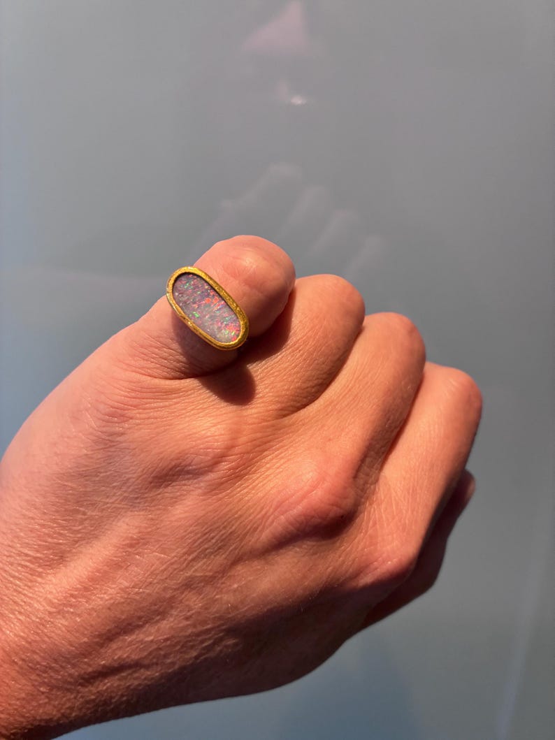 May include: An oval-shaped ring featuring a bezel-set opal gemstone with iridescent flashes of color. The stone is encased in a smooth, polished yellow gold band that follows the elongated oval contour of the central setting.