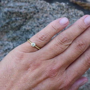 May include: A delicate gold ring with a single, round, clear gemstone set in a bezel. The ring is on a finger, showcasing its simple elegance. The background is a blurred, textured surface.