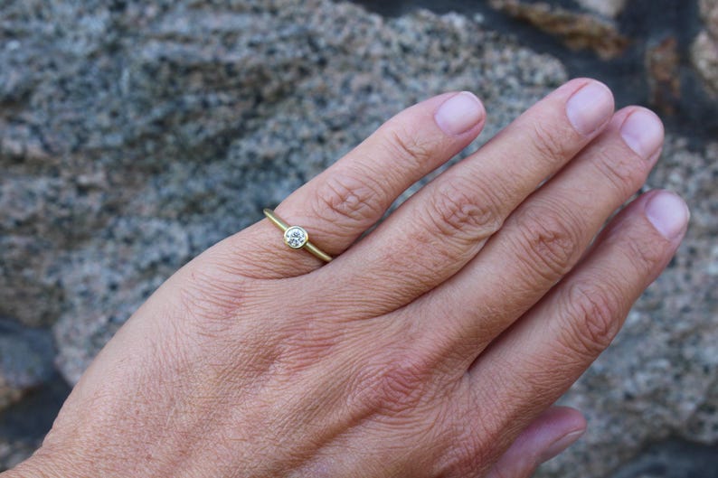May include: A delicate gold ring with a single, round, clear gemstone set in a bezel. The ring is on a finger, showcasing its simple elegance. The background is a blurred, textured surface.