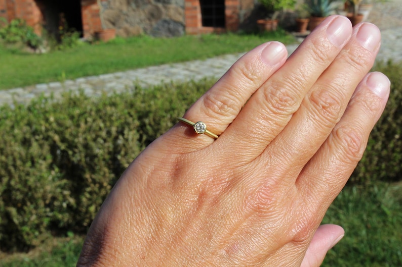 May include: A gold ring with a small, round, clear gemstone set in the center. The ring is on a person's finger, with a green and brown background.
