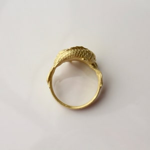 May include: A gold-colored ring with a detailed design. The ring features a textured, possibly scaled, central element and stylized wing-like extensions on either side of the band. The ring is set against a white background.