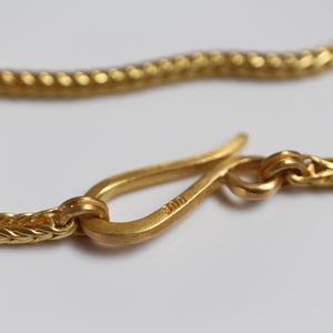 May include: A gold-colored chain necklace featuring a hook-style clasp. The metal surface has a matte finish, and the clasp is clearly stamped with the number 900. The chain links are woven in a tight, repeating pattern that creates a textured appearance.