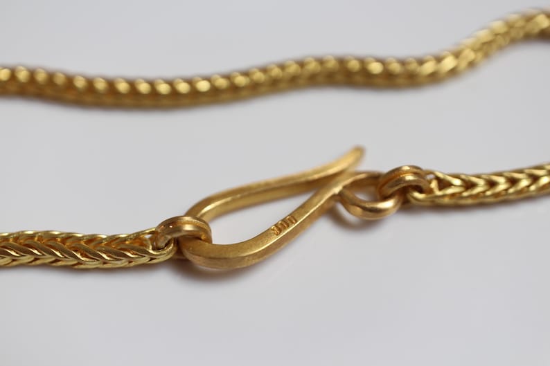 May include: A gold-colored chain necklace featuring a hook-style clasp. The metal surface has a matte finish, and the clasp is clearly stamped with the number 900. The chain links are woven in a tight, repeating pattern that creates a textured appearance.