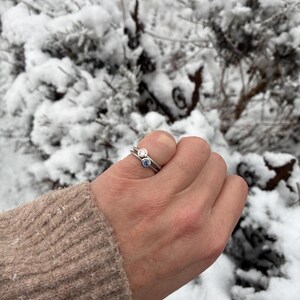 May include: Two silver-toned rings stacked together on a finger. The top ring features a clear, round-cut stone in a bezel setting, while the bottom ring features a light blue, round-cut stone in a matching bezel setting.