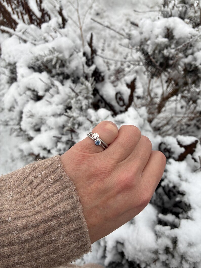 May include: Two silver-toned rings stacked together on a finger. The top ring features a clear, round-cut stone in a bezel setting, while the bottom ring features a light blue, round-cut stone in a matching bezel setting.