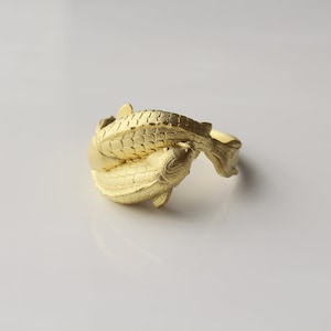 May include: A gold-colored ring featuring a design of two koi fish intertwined. The fish have detailed scales and fins, creating a textured surface. The ring is set against a white background.