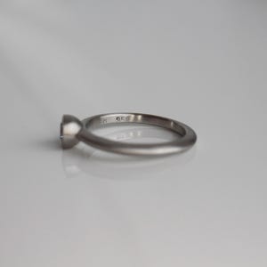 May include: A silver ring with a round, clear gemstone. The ring has a simple, elegant design with a smooth band. The gemstone is set in a bezel setting, and the ring is displayed on a white surface.