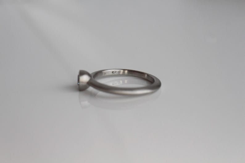 May include: A silver ring with a round, clear gemstone. The ring has a simple, elegant design with a smooth band. The gemstone is set in a bezel setting, and the ring is displayed on a white surface.