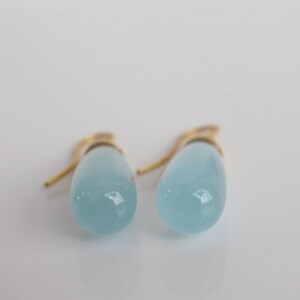 May include: A pair of light blue teardrop-shaped earrings with gold-colored hooks. The earrings have a smooth, glossy finish and are set against a white background. The jewelry is simple and elegant.