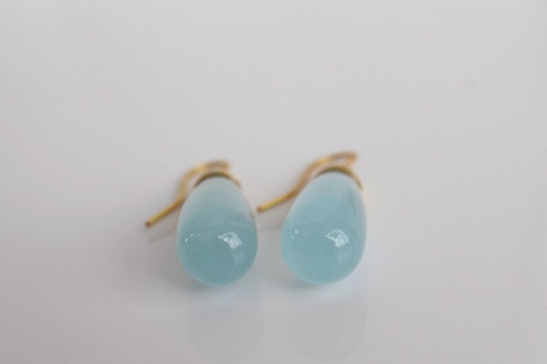 May include: A pair of light blue teardrop-shaped earrings with gold-colored hooks. The earrings have a smooth, glossy finish and are set against a white background. The jewelry is simple and elegant.