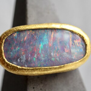 May include: An oval-shaped ring featuring a large, iridescent dark opal gemstone with flashes of blue, green, and red. The stone is set within a thick, textured, matte yellow gold bezel that follows the rounded contour of the gemstone.