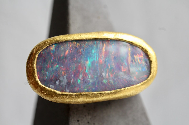 May include: An oval-shaped ring featuring a large, iridescent dark opal gemstone with flashes of blue, green, and red. The stone is set within a thick, textured, matte yellow gold bezel that follows the rounded contour of the gemstone.