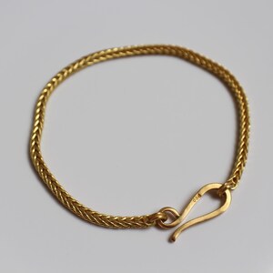 May include: A gold-toned bracelet featuring a woven, herringbone-style chain link design. The bracelet is secured with a curved, hook-shaped clasp mechanism that connects to a circular loop, creating a continuous, flexible circular band.