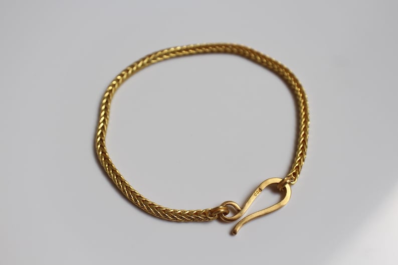 May include: A gold-toned bracelet featuring a woven, herringbone-style chain link design. The bracelet is secured with a curved, hook-shaped clasp mechanism that connects to a circular loop, creating a continuous, flexible circular band.