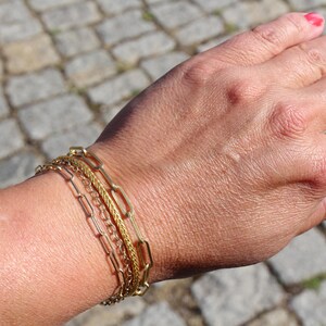 May include: A set of three gold-toned metal bracelets worn together. The set includes one bracelet with elongated rectangular links, one with a fine twisted rope texture, and one with a delicate, thin chain design, all featuring a polished finish.