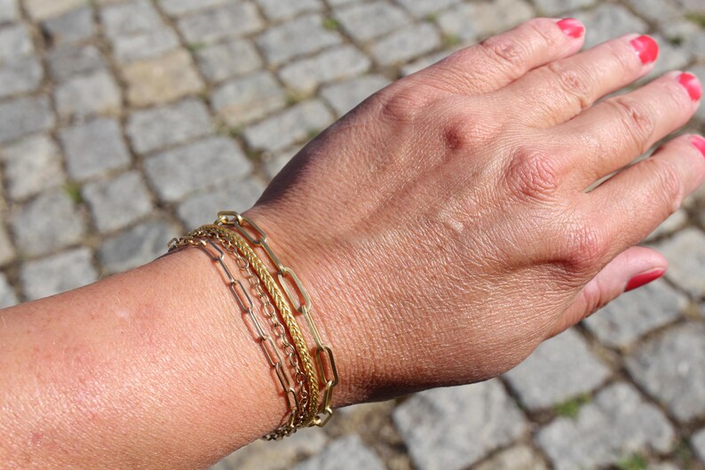 May include: A set of three gold-toned metal bracelets worn together. The set includes one bracelet with elongated rectangular links, one with a fine twisted rope texture, and one with a delicate, thin chain design, all featuring a polished finish.