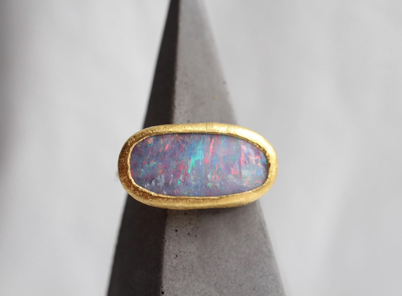 May include: An oval-shaped ring featuring a dark, iridescent opal gemstone with flashes of red, blue, and green light. The stone is set within a textured, matte yellow gold bezel that follows the elongated horizontal shape of the opal.