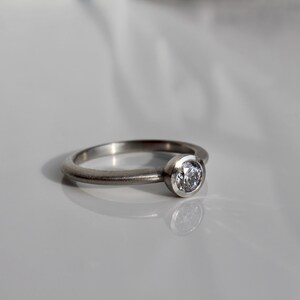 May include: A silver-toned metal ring featuring a single round, clear, faceted stone set in a bezel setting. The band has a smooth, matte finish and a rounded profile, reflecting light on its surface while holding the central gemstone.