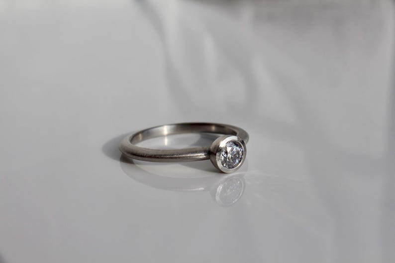 May include: A silver-toned metal ring featuring a single round, clear, faceted stone set in a bezel setting. The band has a smooth, matte finish and a rounded profile, reflecting light on its surface while holding the central gemstone.