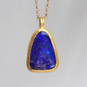 Könnte beinhalten: Eine Goldhalskette mit einem dreieckigen, tiefblauen Opal-Anhänger mit irisierenden Sprenkeln. Der Stein ist in eine strukturierte, matte Goldfassung eingefasst und hängt an einer zarten Goldkette mit kleinen, runden, miteinander verbundenen Gliedern.